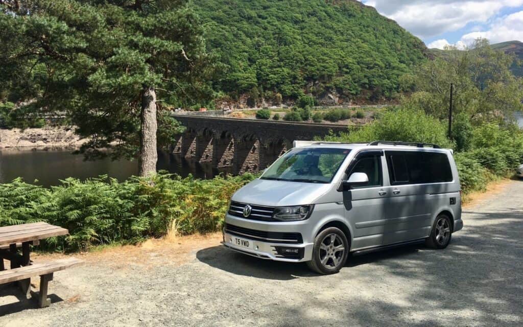 Silver Volkswagen camper van parked near a river with lush green trees and mountains in the backgrou.